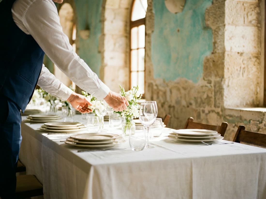 A long table being laid with white linen, stacked plates, glasses and white flowers in a stone hall